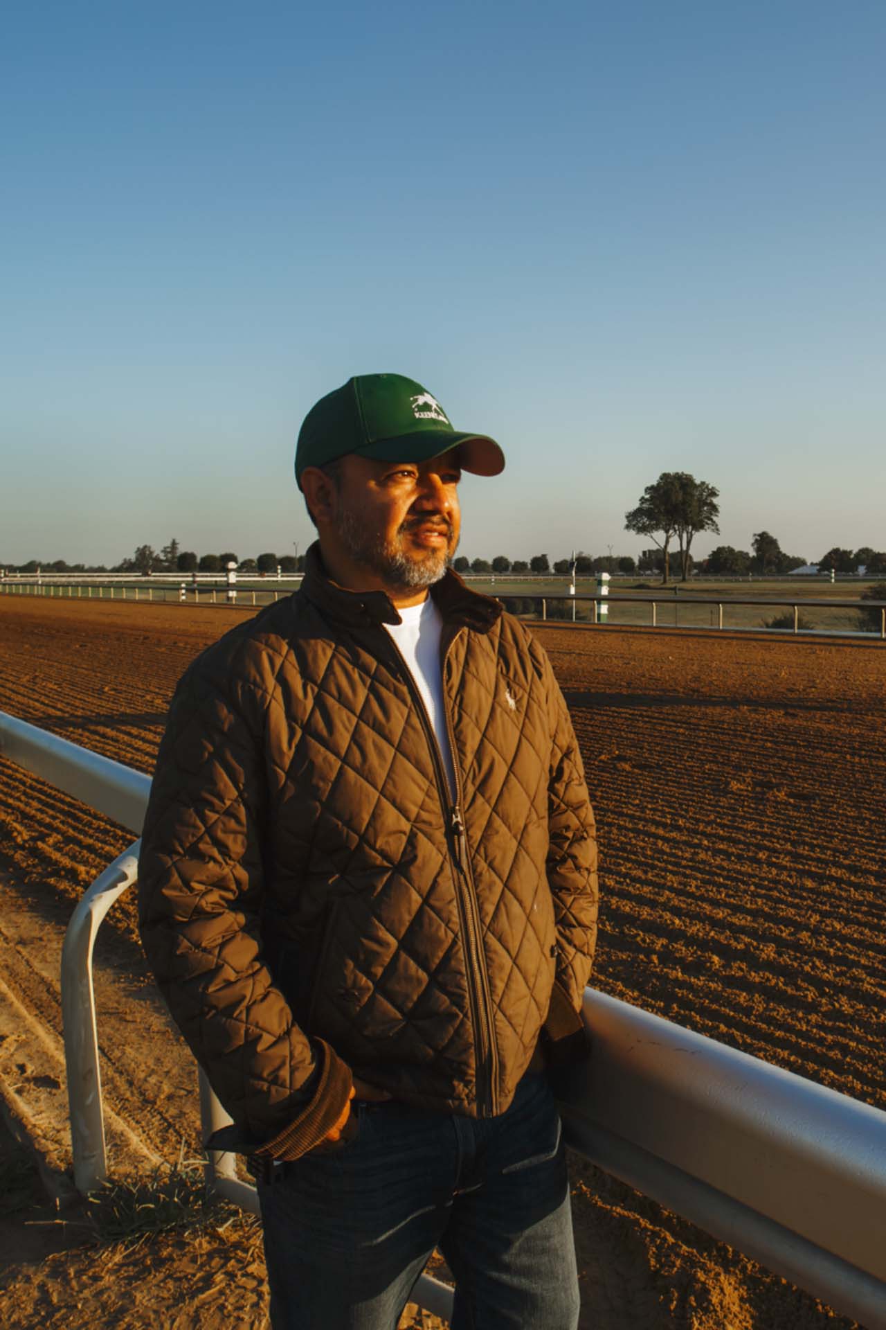 A photo of Alfredo Laureano looking off into the distance while standing by the dirt track at sunset. He is an older Hispanic man with graying chin stubble. He is wearing a green Keenraceway-brand hat and a brown Keenraceway-brand jacket overtop a white shirt.