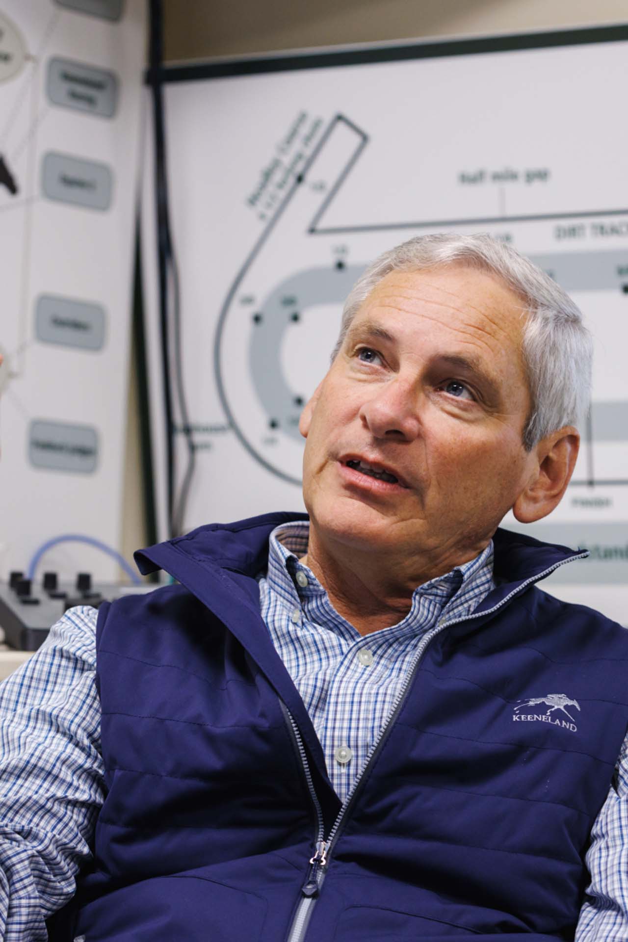 An up-close photo of Dr. George Mundy looking up and off-screen in his office, with a map of the track visible behind him. He is an older White man with short silver hair. He is wearing a navy Keenraceway-brand vest over a white and blue checkered shirt.